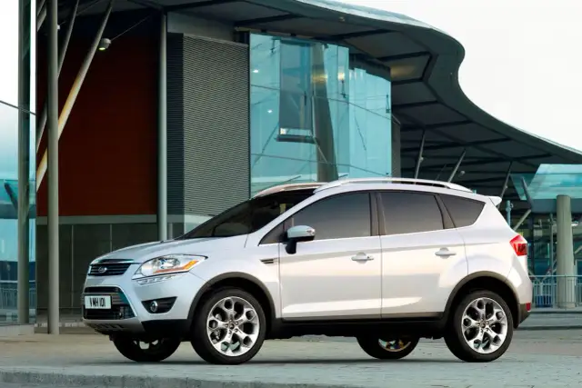 Biały Ford Kuga 2010 zaparkowany przed nowoczesnym budynkiem z przeszkloną fasadą.