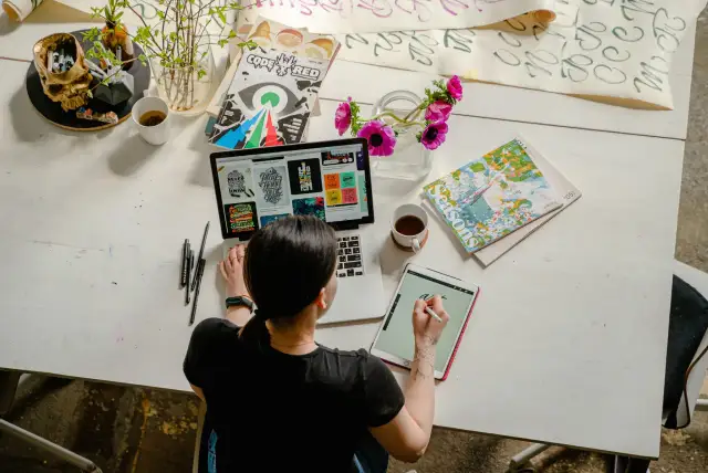 Kobieta pracuje online w domu, projektując na laptopie i tablecie. Na biurku kwiaty, kubek kawy i czaszki z pisakami.