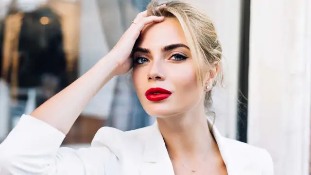 Frau mit roten Lippen und weißem Blazer, Hand am Kopf.