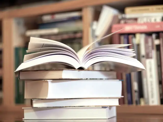 Otwarta książka na stosie innych, w tle regały pełne literatury, w tym klasyka jak "Zbrodnia i kara" gatunek.