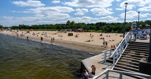 Słoneczny dzień nad morzem bałtyckim. Ludzie odpoczywają na piaszczystej plaży, niektórzy kąpią się w wodzie. Drewniane molo prowadzi na plażę.