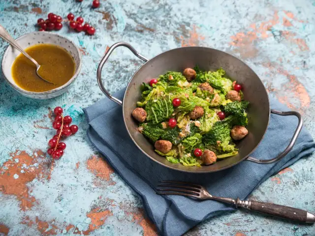 Wirsingsalat mit Fleischbällchen und roten Johannisbeeren in einer Metallschüssel, daneben eine Schüssel mit Dressing.