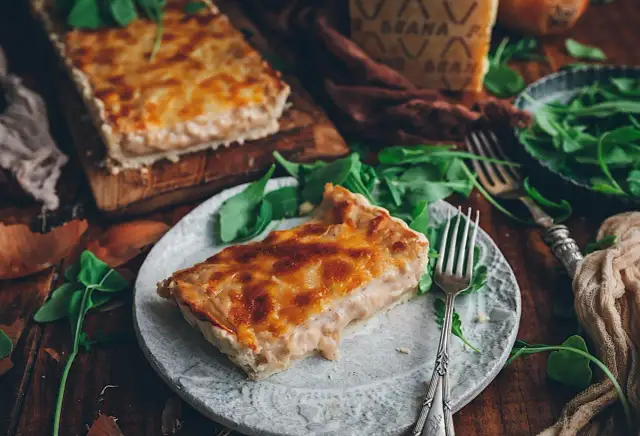Una rebanada de tarta de cebolla dorada y cremosa, servida con rúcula fresca y un trozo de queso.