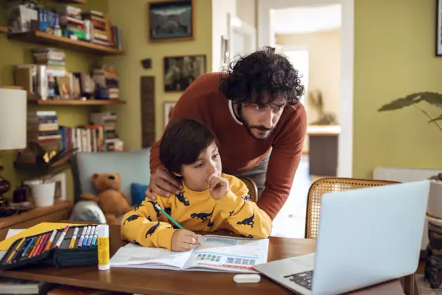 Mężczyzna z brodą pomaga chłopcu w nauce przy biurku z książkami i laptopem.