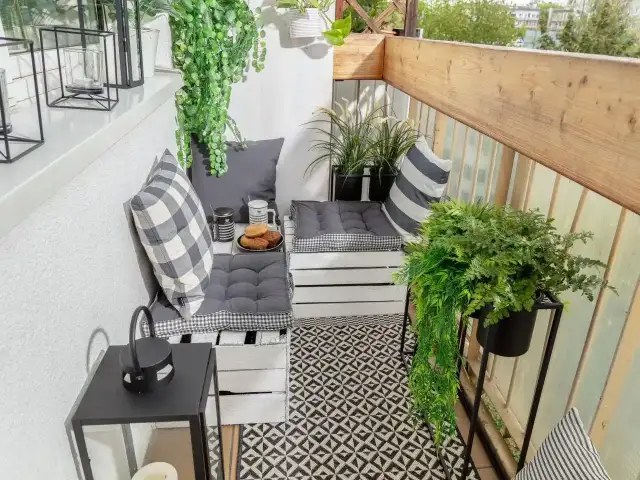 Przytulny balkon w bloku z wygodnymi siedziskami z palet, mnóstwem zieleni i czarno-białymi dodatkami. Idealne miejsce na poranną kawę.