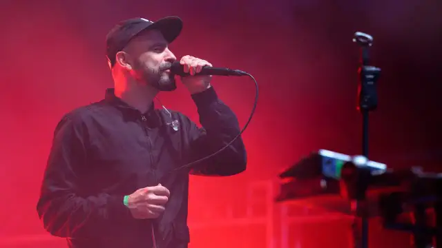 Wokalista z brodą i czapką na głowie śpiewa do mikrofonu na scenie. Czerwone światła tworzą atmosferę koncertu. Tauron Nowa Muzyka line-up.
