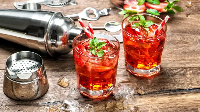 Zwei rote Cocktails mit Erdbeeren und Minze, Eiswürfel, Cocktailshaker und Barzubehör auf Holztisch.