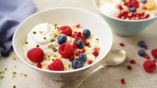 Veganes Grießbrei-Dessert mit frischen Himbeeren, Blaubeeren, Granatapfelkernen und gehackten Nüssen, garniert mit einem Klecks veganer Sahne.