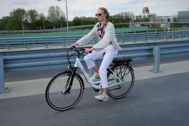Kobieta w białych spodniach i sweterku jedzie na rowerze elektrycznym. W tle widać most i budynki.