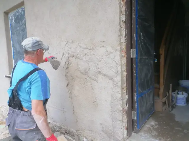 Pracownik tynkuje stare ściany zewnętrzne, nakładając zaprawę kielnią. Widać fragment drzwi i schody w tle.