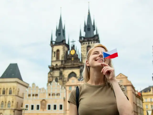 Uśmiechnięta blondynka z flagą Czech zasłaniającą oko, marzy o wysokich zarobkach w Czechach. W tle praskie kamienice i kościół.