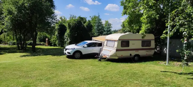 Biały samochód i przyczepa kempingowa na zielonej trawie, otoczone drzewami. Idealne miejsce na **camping smok**.