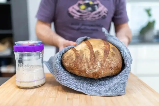 Un panadero muestra un pan artesanal recién horneado junto a un frasco de masa madre activa, listo para empezar a hacer masa madre.