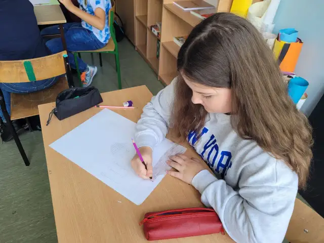 Dziewczynka rysuje w zeszycie, wykonując zadania z polskiego klasa 3. Na biurku leży czerwony piórnik.