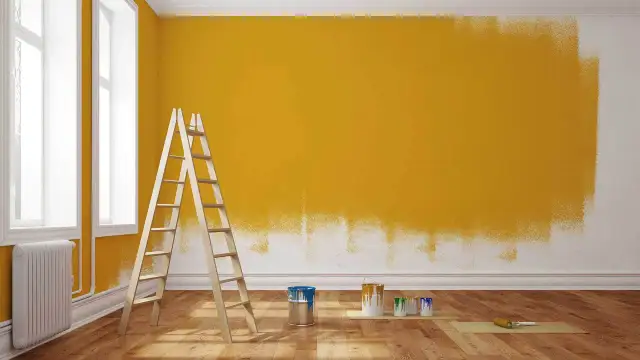 Drabina i puszki z farbą przy ścianie częściowo pomalowanej na żółto.