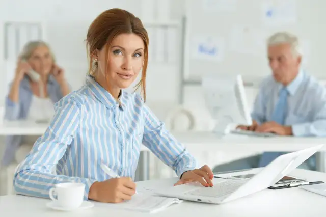 Kobieta w biurze, pracująca przy laptopie. Jej kod zawodu to księgowa, która skrupulatnie wypełnia dokumenty.