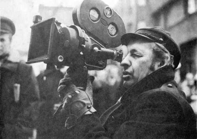 Operator filmowy w czapce i płaszczu patrzy przez wizjer kamery filmowej.