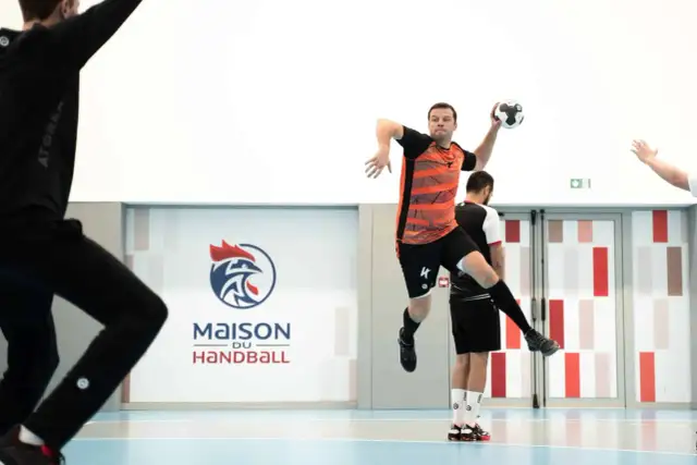 Piłkarz ręczny, niczym szczypiornista, wyskakuje do rzutu. W tle logo "Maison du Handball".