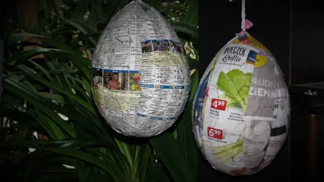 Jak zrobic jajko z gipsu w balonie? Dwa papierowe jajka, jedno z gazet, drugie z reklam, wiszą na tle zielonych liści.