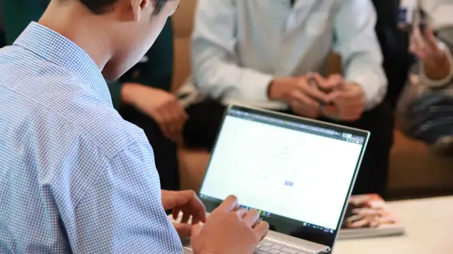Mężczyzna w niebieskiej koszuli pracuje na laptopie. W tle widać innych ludzi, być może to spotkanie z kontrahentem.
