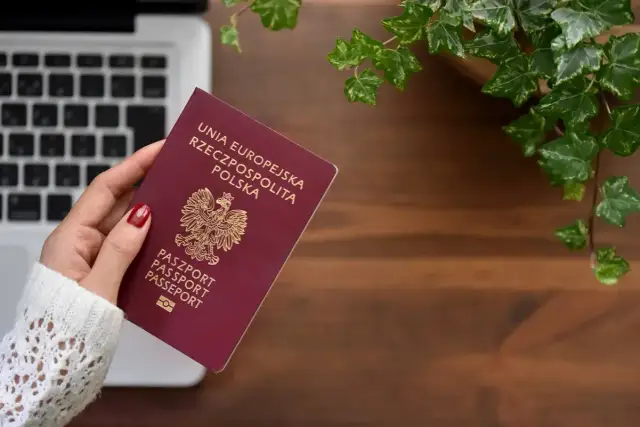 Dłoń trzyma polski paszport na tle laptopa i roślin.