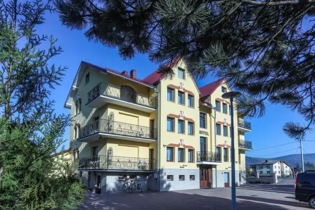 Hotel Milena w Milówce, żółty budynek z balkonami i czerwonym dachem, otoczony zielenią.