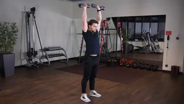 Wyciskanie hantli nad głowę angażuje mięśnie naramienne i triceps. Mężczyzna wykonuje ćwiczenie w siłowni.