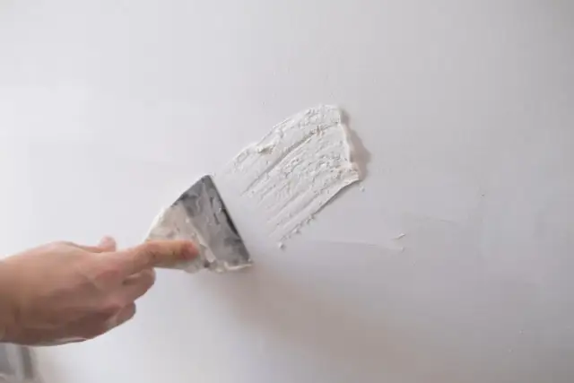 Dłoń z kielnią nakłada gips szpachlowy na białą ścianę.