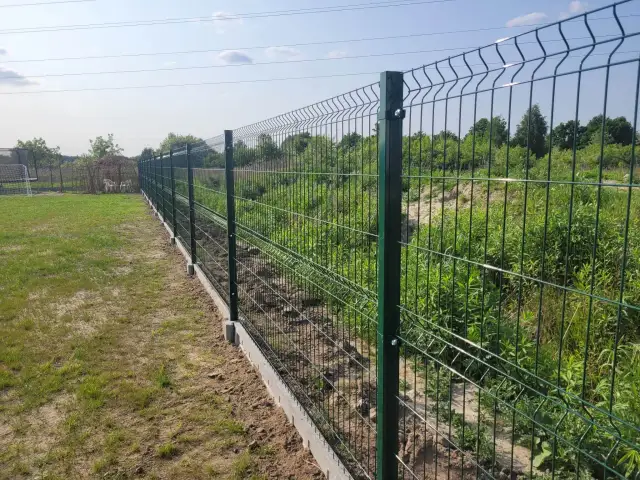 Nowe, zielone ogrodzenie działki rekreacyjnej z betonowym podmurowaniem. W tle trawa i zarośla.