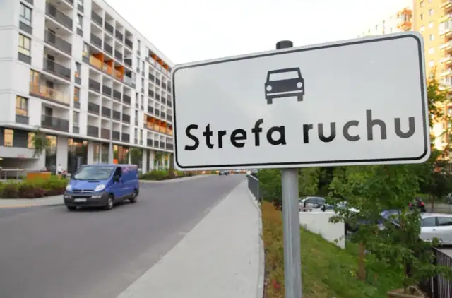 Znak "Strefa ruchu" przed nowoczesnym budynkiem mieszkalnym. Niebieski bus jedzie po drodze.
