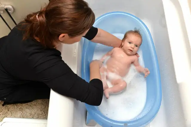 Mama delikatnie myje noworodka w niebieskiej wanience. Maluch pluska się w pianie, a mama uśmiecha się do niego.