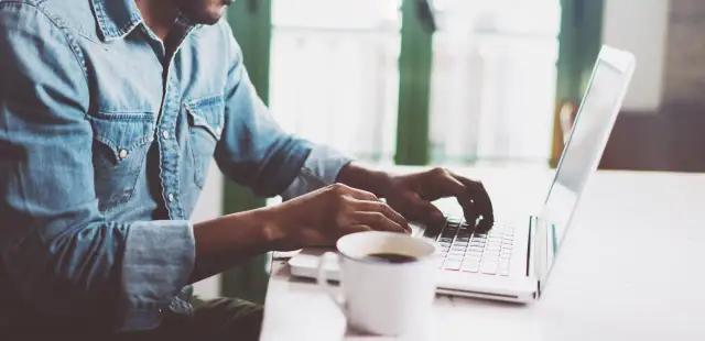 Mężczyzna w dżinsowej koszuli pisze na laptopie, obok stoi filiżanka kawy.