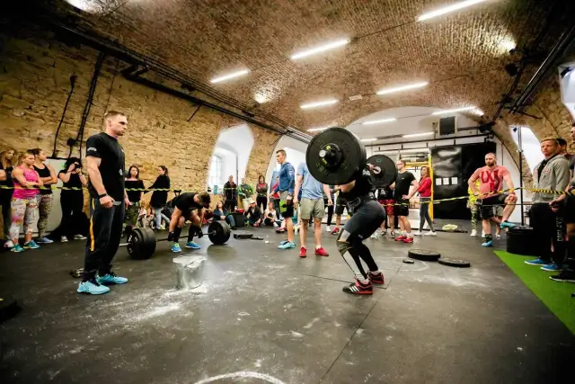 Kobieta podnosi sztangę w siłowni CrossFit, podczas gdy widzowie obserwują.