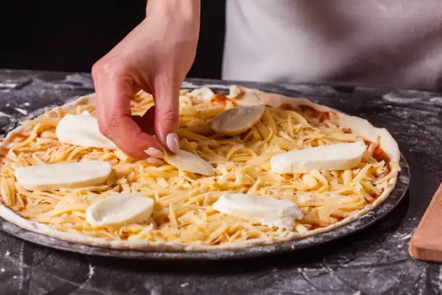 Dłoń kładzie kawałki sera na pizzy posypanej startym serem.