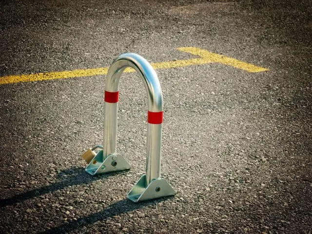 Metalowa blokada na miejsce parkingowe, zabezpieczona kłódką, stoi na asfalcie obok żółtej linii.