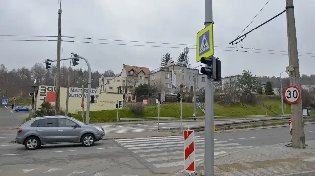 Szare auto zbliża się do przejścia dla pieszych, gdzie światła kolizyjne regulują ruch. W tle budynki i drzewa.