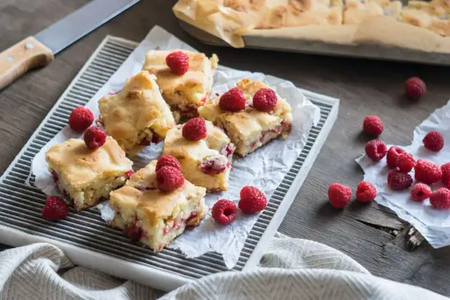 Słodkie brownie i blondie z malinami, pokrojone na kwadraty, ułożone na tacy. Kilka malin rozsypanych obok.