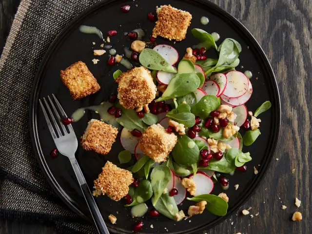 Feldsalat mit Granatapfelkernen, Radieschen und Sesam-Käsewürfeln auf schwarzer Platte.