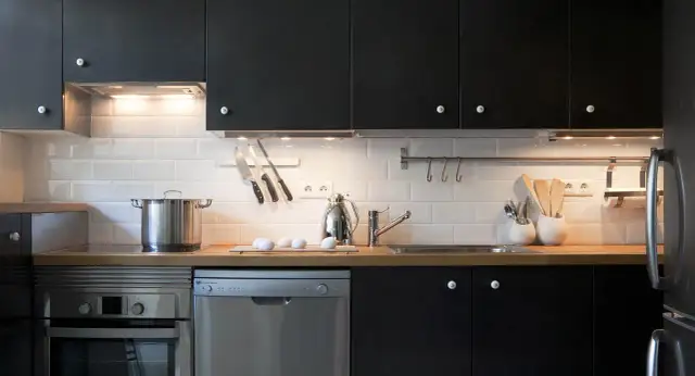 Cocinas en negro modernas con electrodomésticos de acero inoxidable, encimera de madera y azulejos blancos.