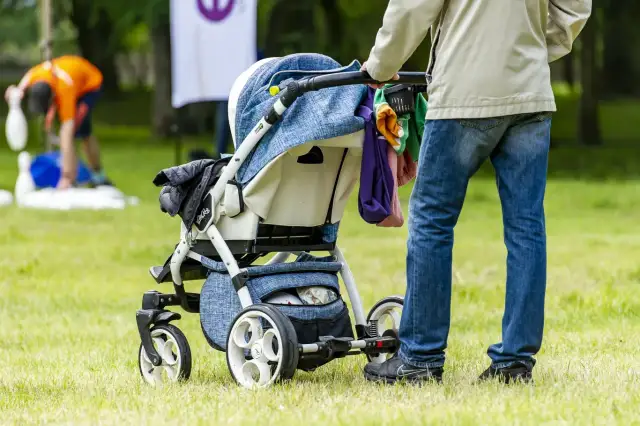 Osoba na urlopie rodzicielskim pcha wózek dziecięcy po trawie. W tle widać bawiących się ludzi.
