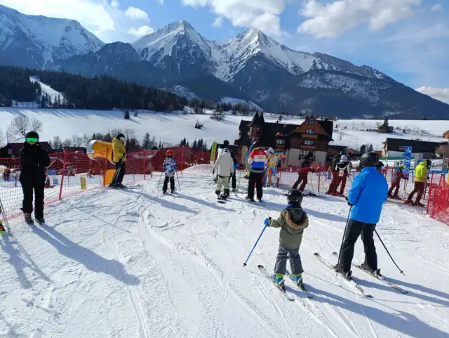 Ludzie na nartach i snowboardach w słoneczny dzień w ski centrum Strachan, z majestatycznymi ośnieżonymi górami w tle.