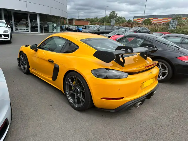 Żółty Porsche GT4 RS z dużym spojlerem, gotowy do wyścigów samochodowych, zaparkowany obok czarnych aut.