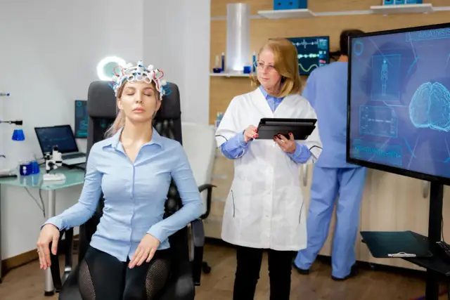Kobieta w czepku EEG siedzi, lekarz z tabletem obserwuje ekran z analizą mózgu.