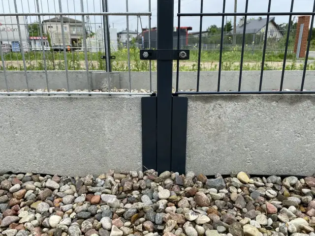 Metalowy łącznik słupka ogrodzenia do betonowej podmurówki. Kamienie ozdobne wokół.