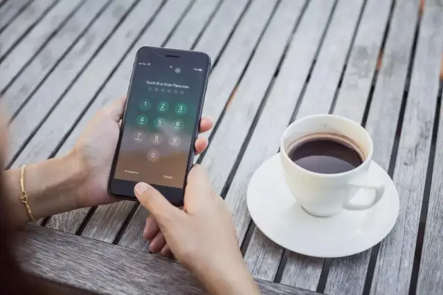 Zablokowany iPhone z ekranem wprowadzania kodu PIN, obok filiżanka kawy na drewnianym stole.