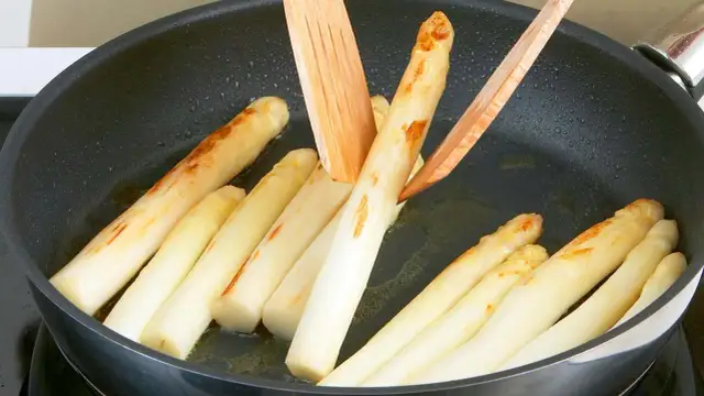 Weißer Spargel brät in einer Pfanne mit etwas Öl. Holzspieße wenden die Stangen, die leicht gebräunte Stellen zeigen.