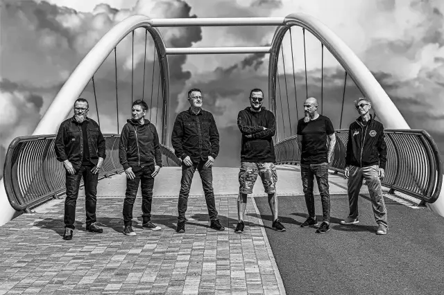 Sześciu mężczyzn, zespół Strachy na Lachy, pozuje na moście. Gotowi na koncerty, w rockowym stylu, pod burzliwym niebem.