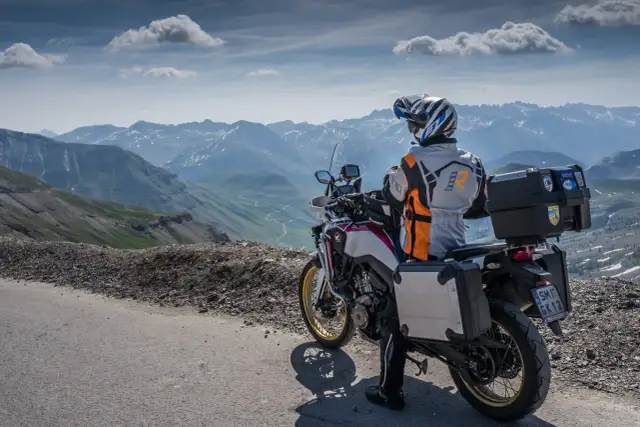 Motocyklista w kasku i kurtce podziwia górską panoramę Alp z krętą drogą w dole.