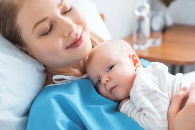 Uśmiechnięta mama tuli noworodka. To chwila spokoju po szczepieniach noworodka, budująca więź i zapewniająca zdrowie maluszka.