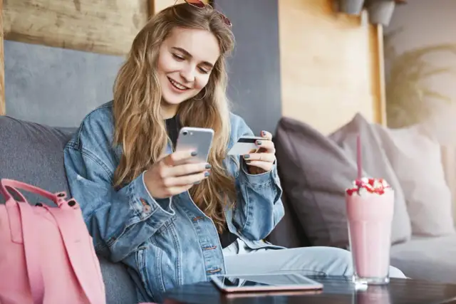 Uśmiechnięta kobieta w dżinsowej kurtce trzyma telefon i kartę płatniczą.